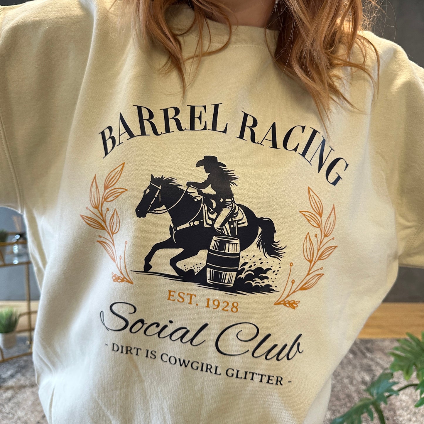 Design on a sand-colored crewneck featuring a navy blue silhouette of a cowgirl barrel racing on horseback, encircled by elegant golden leaf accents. The text reads 'BARREL RACING Social Club' in stylized navy blue and gold lettering, with 'EST. 1928' in gold beneath the illustration. Below, in smaller text, it says '- Dirt is Cowgirl Glitter -'.