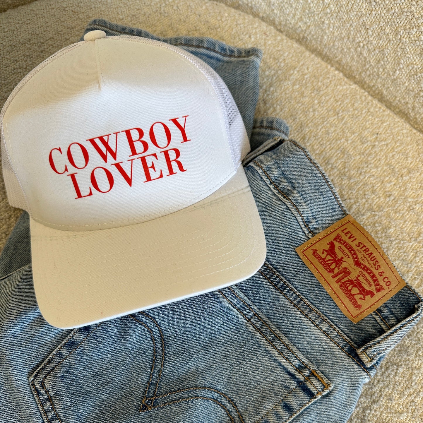 A white trucker hat featuring the 'COWBOY LOVER' logo, in red. The item is displayed on a beige textured surface next to a pair of Levi Strauss 501 Originals jeans. Creating a fun and stylish western-inspired look.