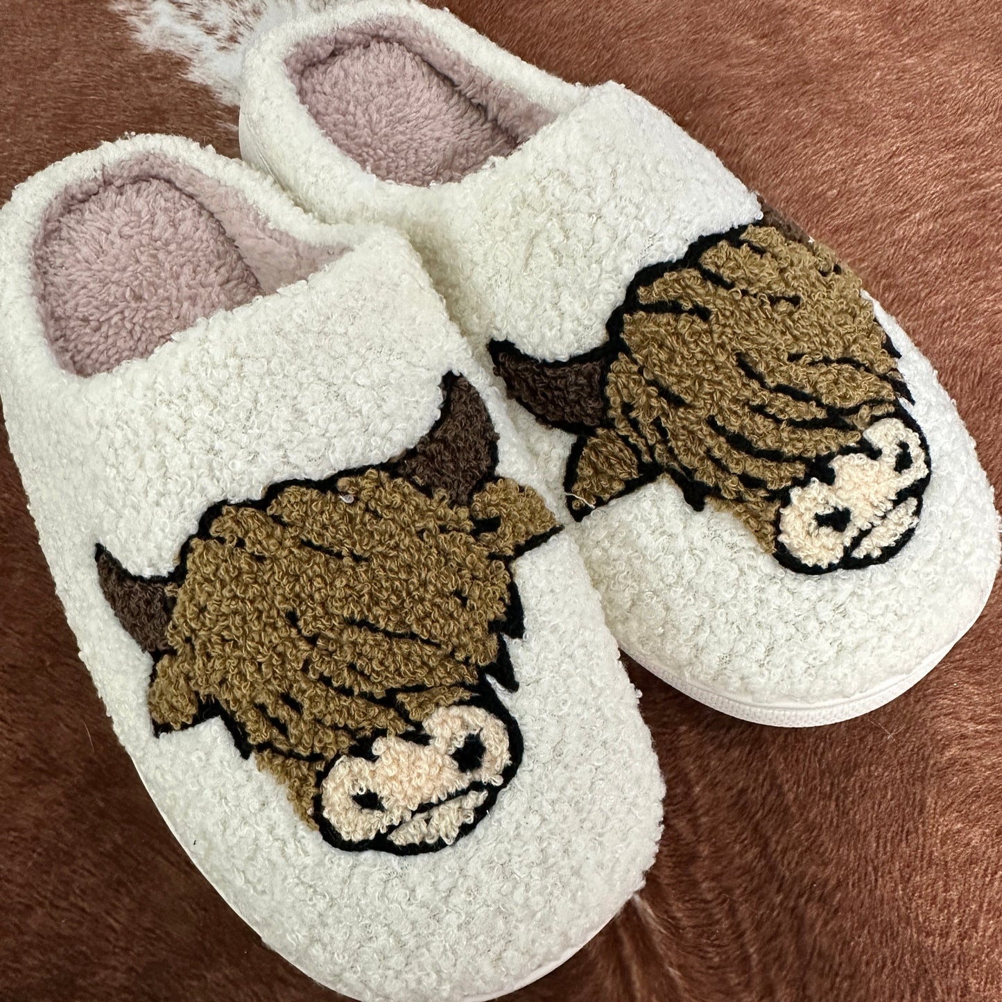 A cozy pair of cream-colored, sherpa-style slippers with an adorable Highland cow design embroidered on the front. The cows have fluffy brown fur, small horns, and a cute snout, giving them a charming rustic vibe. The slippers are placed on a cowhide rug, adding to the Western aesthetic.