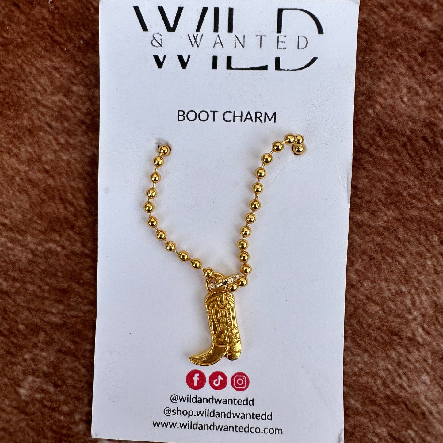 Gold boot charm on a card with 'Wild & Wanted' branding on a brown textured background