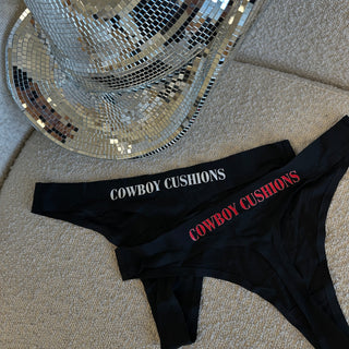 Set of two black thong underwear featuring the 'COWBOY CUSHIONS' logo, one with white lettering and the other with red. The items are displayed on a beige textured surface next to a silver disco ball cowboy hat, creating a fun and stylish western-inspired look.