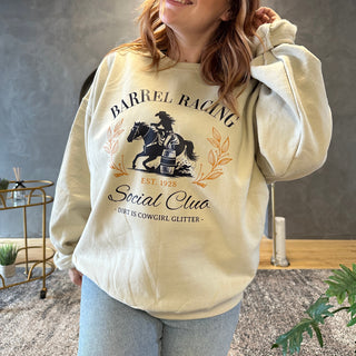 Design on a sand-colored crewneck featuring a navy blue silhouette of a cowgirl barrel racing on horseback, encircled by elegant golden leaf accents. The text reads 'BARREL RACING Social Club' in stylized navy blue and gold lettering, with 'EST. 1928' in gold beneath the illustration. Below, in smaller text, it says '- Dirt is Cowgirl Glitter -'.