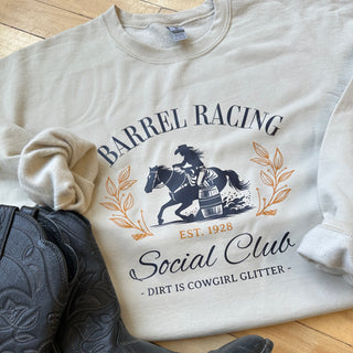 Design on a sand-colored crewneck featuring a navy blue silhouette of a cowgirl barrel racing on horseback, encircled by elegant golden leaf accents. The text reads 'BARREL RACING Social Club' in stylized navy blue and gold lettering, with 'EST. 1928' in gold beneath the illustration. Below, in smaller text, it says '- Dirt is Cowgirl Glitter -'.