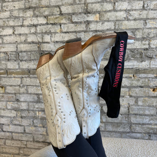 Pair of cream-colored western cowboy boots with intricate embroidered patterns and silver stud detailing. The boots are propped up on a textured stone wall background, with a black strap labeled 'COWBOY CUSHIONS' in bold pink lettering attached to one boot.