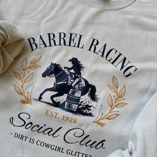 Design on a sand-colored crewneck featuring a navy blue silhouette of a cowgirl barrel racing on horseback, encircled by elegant golden leaf accents. The text reads 'BARREL RACING Social Club' in stylized navy blue and gold lettering, with 'EST. 1928' in gold beneath the illustration. Below, in smaller text, it says '- Dirt is Cowgirl Glitter -'.