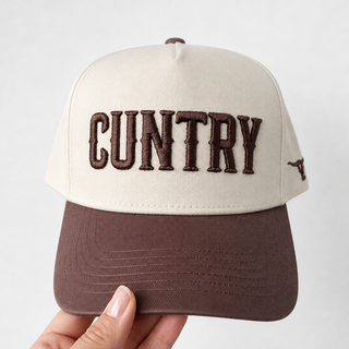 A two-tone baseball cap held by a hand against a light gray background. The cap features an off-white crown with dark brown embroidered text reading 'COUNTRY' and a matching dark brown visor.