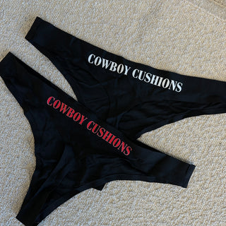 Set of two black thong underwear featuring the 'COWBOY CUSHIONS' logo, one with white lettering and the other with red. The items are displayed on a white textured surface.