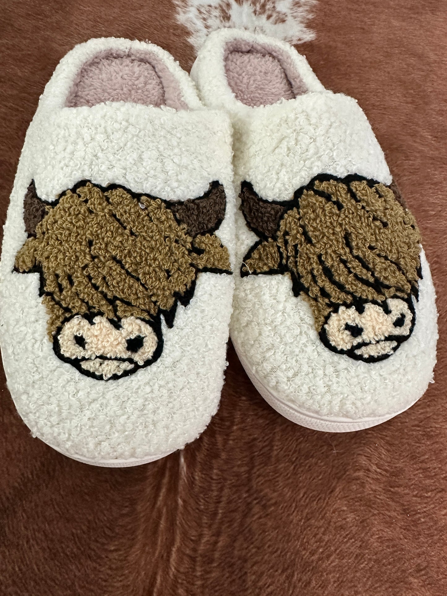 A cozy pair of cream-colored, sherpa-style slippers with an adorable Highland cow design embroidered on the front. The cows have fluffy brown fur, small horns, and a cute snout, giving them a charming rustic vibe. The slippers are placed on a cowhide rug, adding to the Western aesthetic.