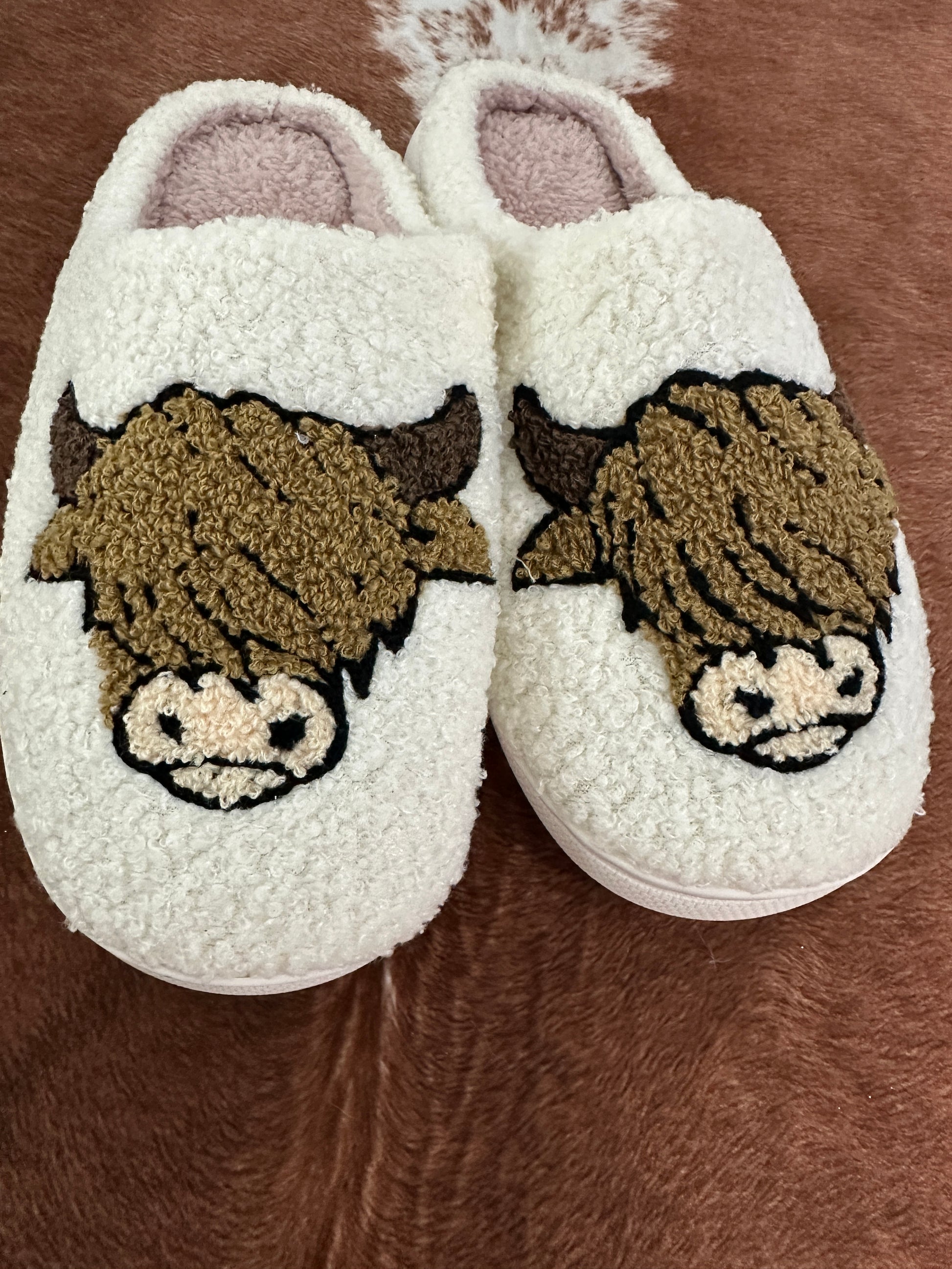 A cozy pair of cream-colored, sherpa-style slippers with an adorable Highland cow design embroidered on the front. The cows have fluffy brown fur, small horns, and a cute snout, giving them a charming rustic vibe. The slippers are placed on a cowhide rug, adding to the Western aesthetic.