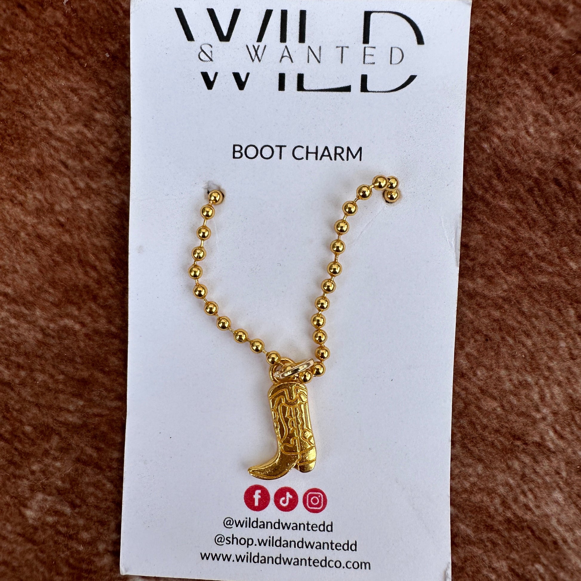 Gold boot charm on a card with 'Wild & Wanted' branding on a brown textured background